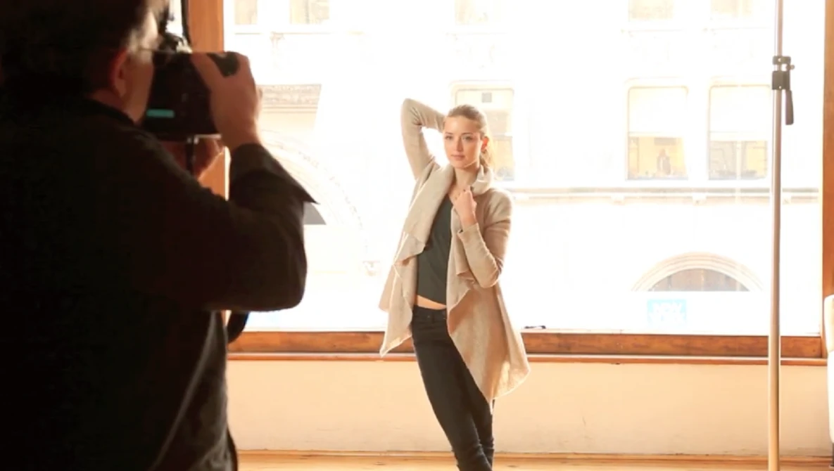 Photographer shooting a portrait of a woman posing in a bright studio with large windows.