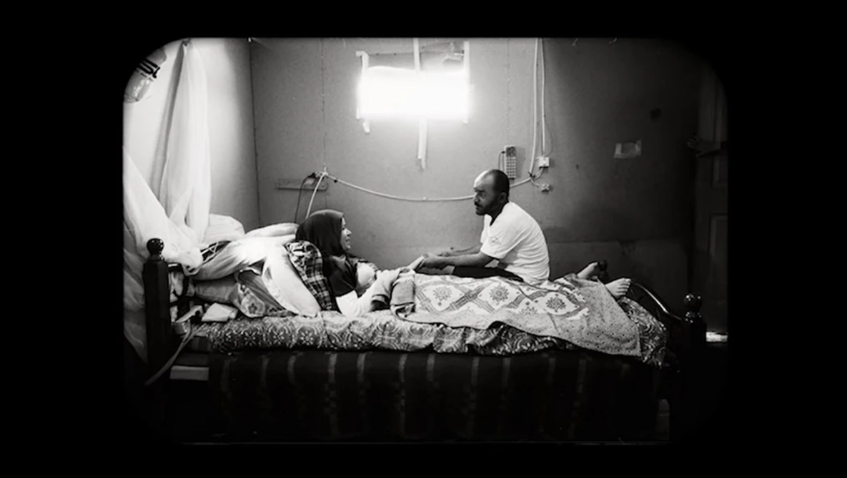 Black and white photograph of two people on a bed in a sparsely furnished room with bright overhead lighting.