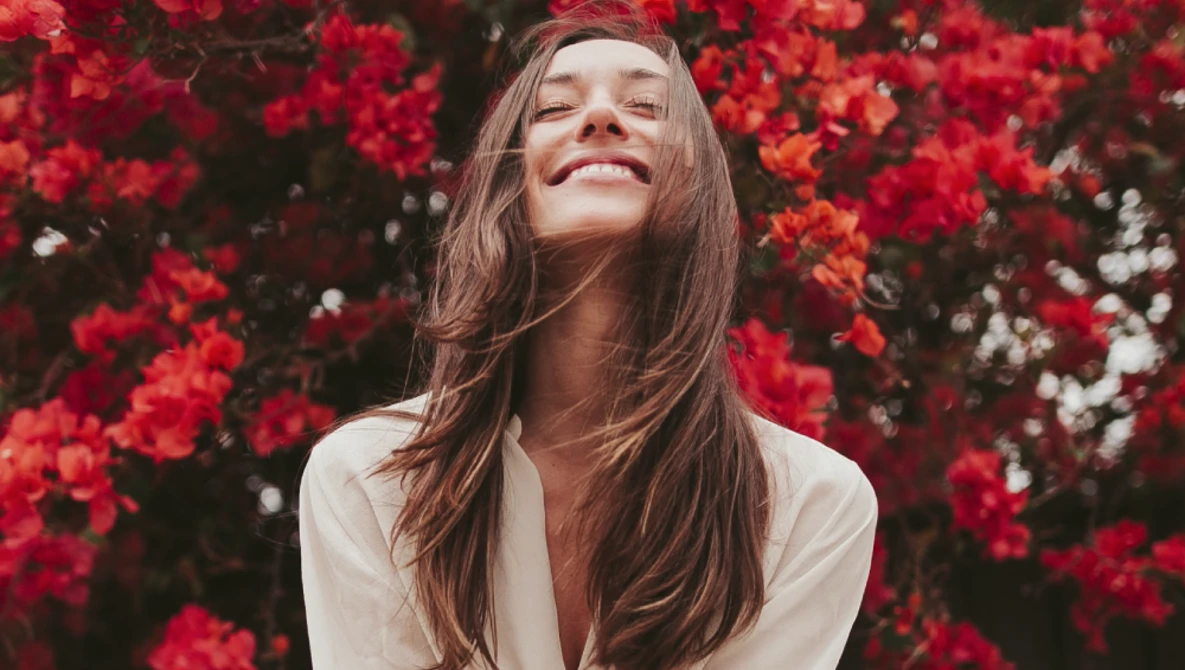 Woman with long brown hair smiling upward among vibrant red flowers in natural sunlight.