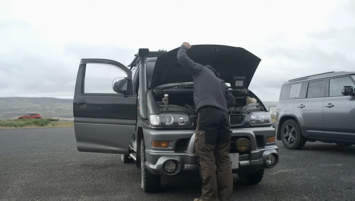 Photographer inspecting engine of parked vehicle during roadside shoot with overcast sky and mountains in background.