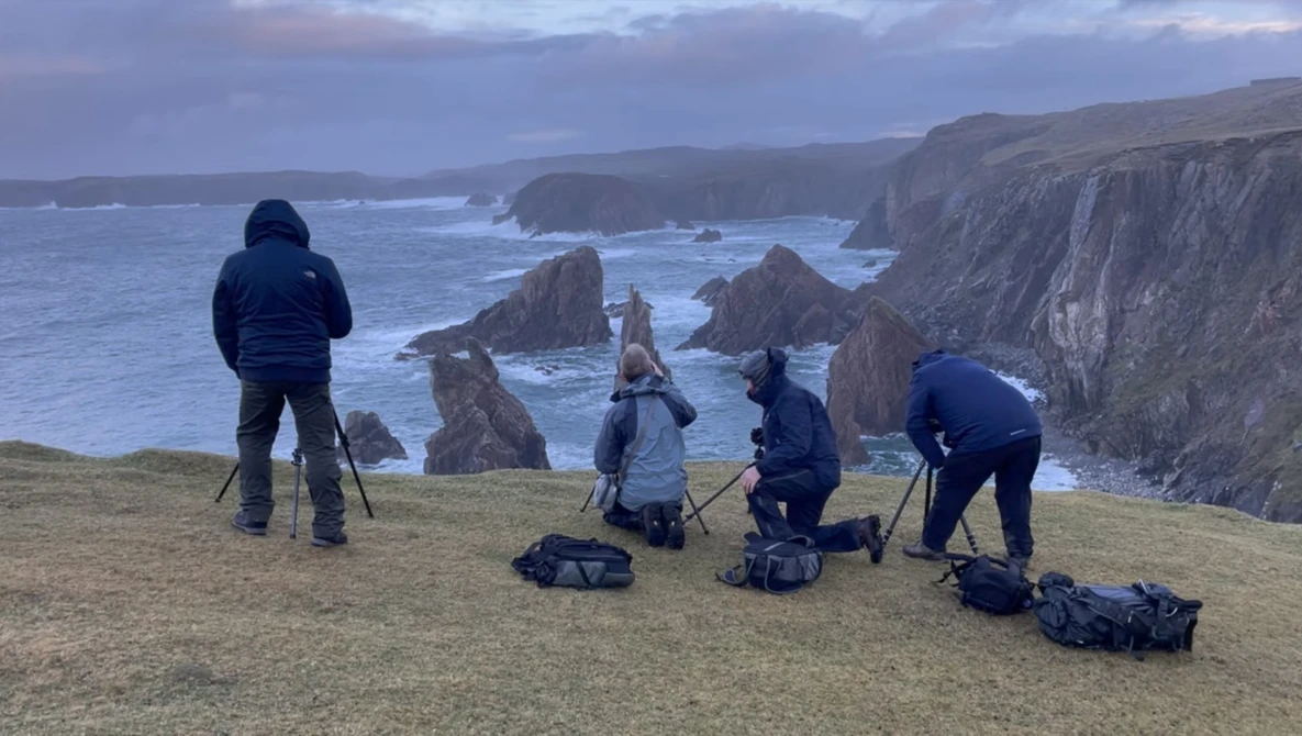 Photography crew of five setting up equipment on a coastal cliff overlooking dramatic rock formations and sea stacks.