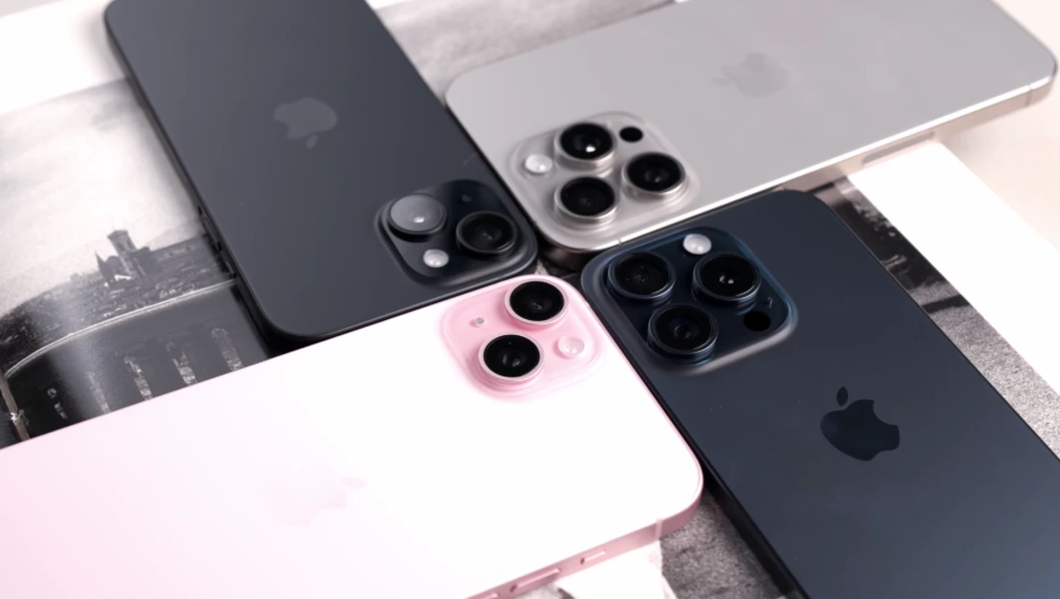 Four iPhones arranged on a white surface, displaying various color options including black, silver, pink, and midnight blue with triple-lens camera systems.