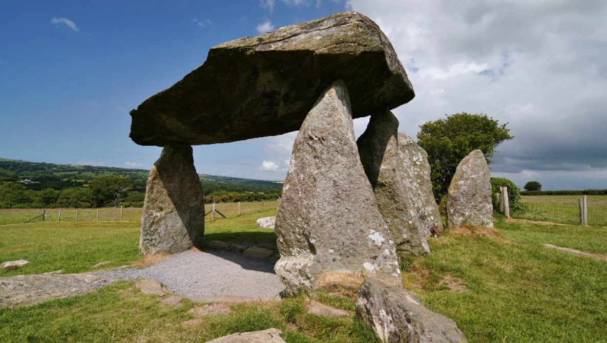 Ancient stone dolmen with large capstone balanced on upright stones in green pastoral landscape.