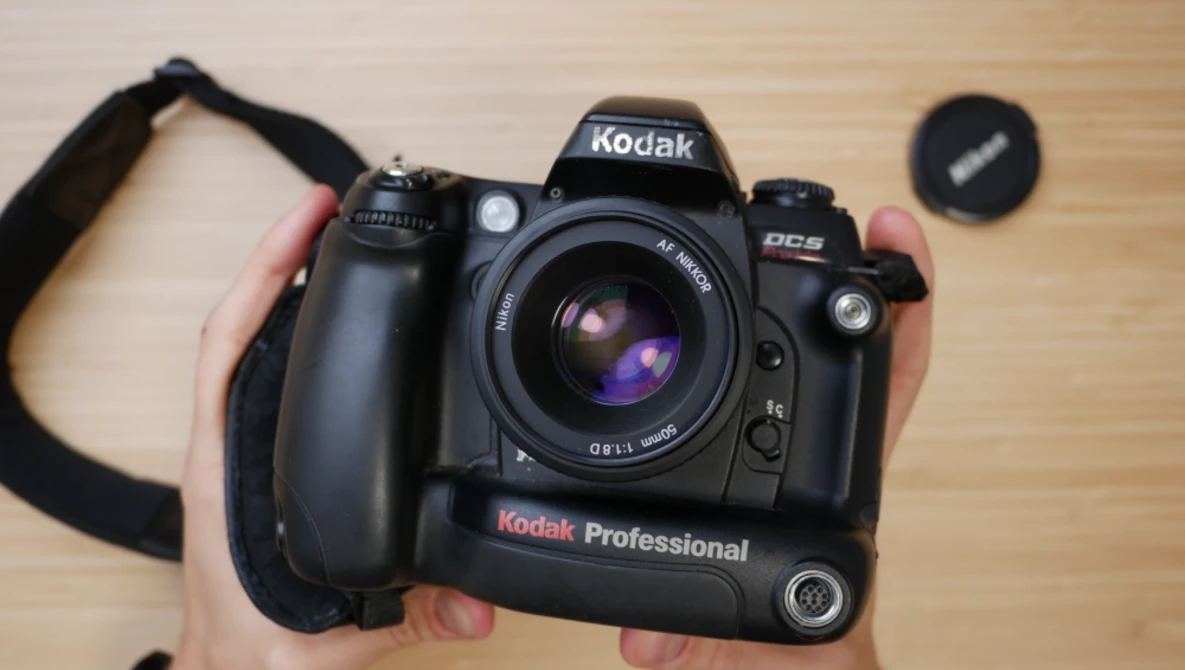 Hands holding a Kodak Professional DCS digital camera with attached grip and lens on wooden surface.