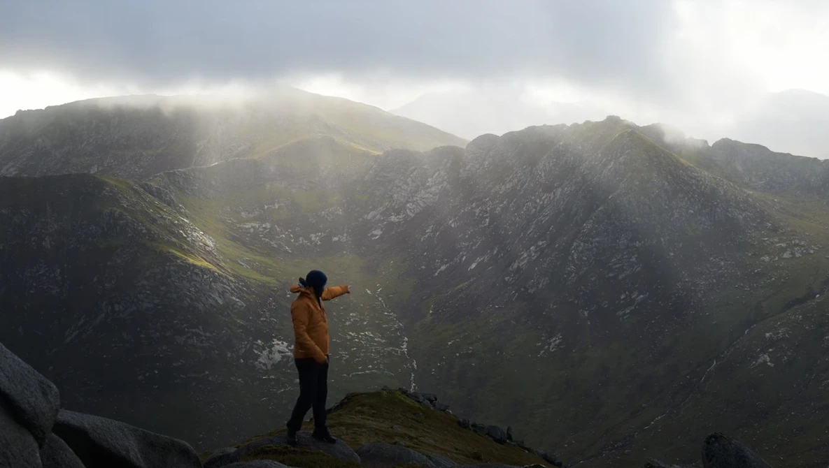 Hiker in yellow jacket standing on rocky ridge overlooking misty mountain valley.