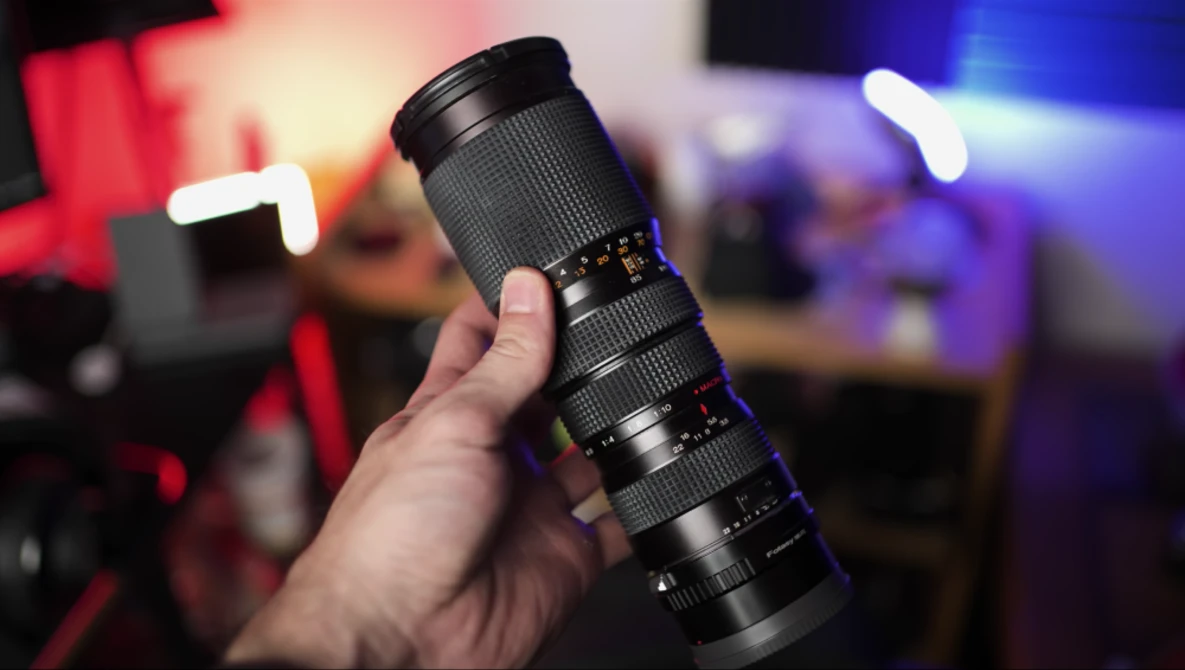 Hand holding a telephoto zoom lens with textured black barrel against blurred colorful background.