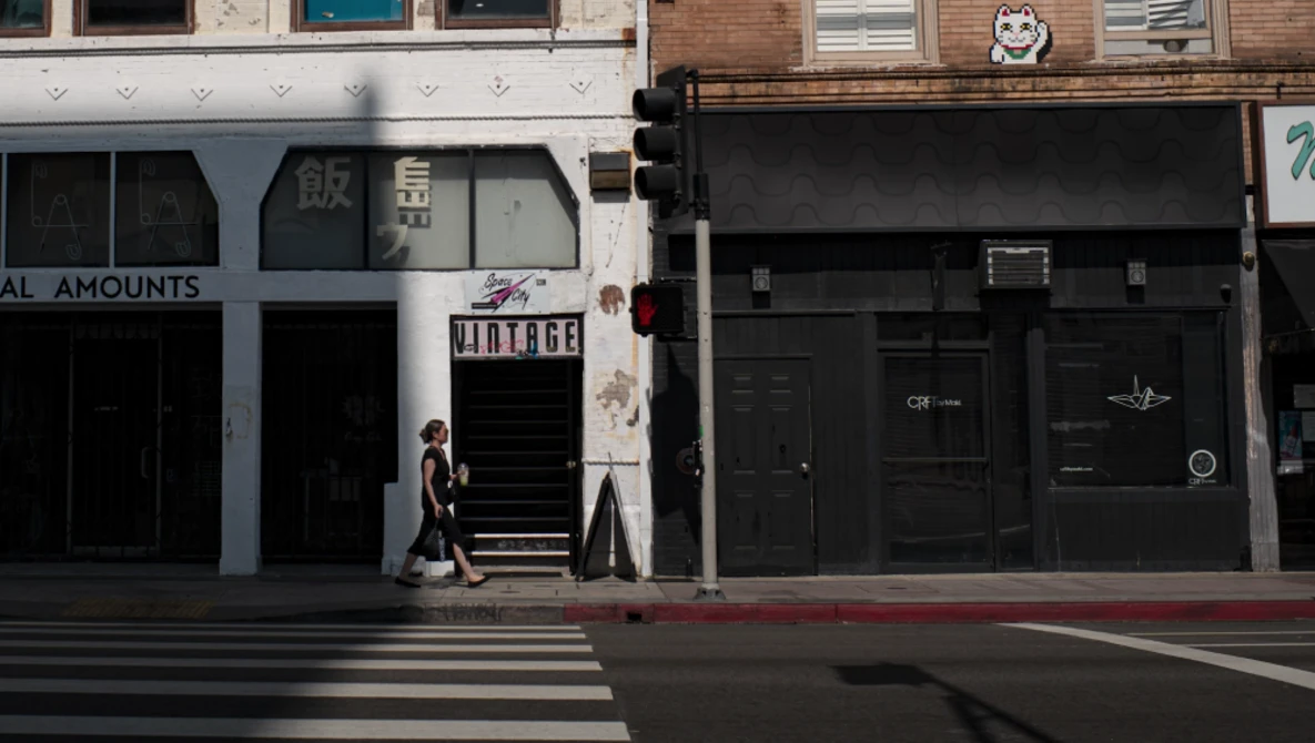 Person walking across crosswalk in front of storefronts with mixed architectural styles and signage.