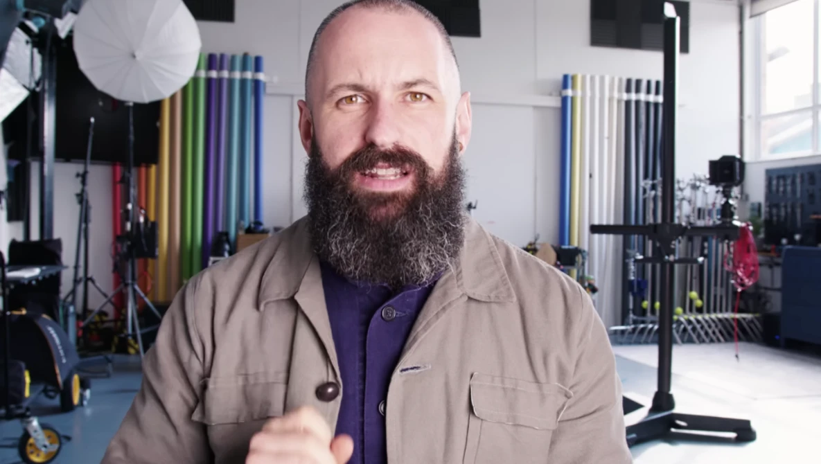 Bearded man in tan jacket gestures while speaking in a photography studio.