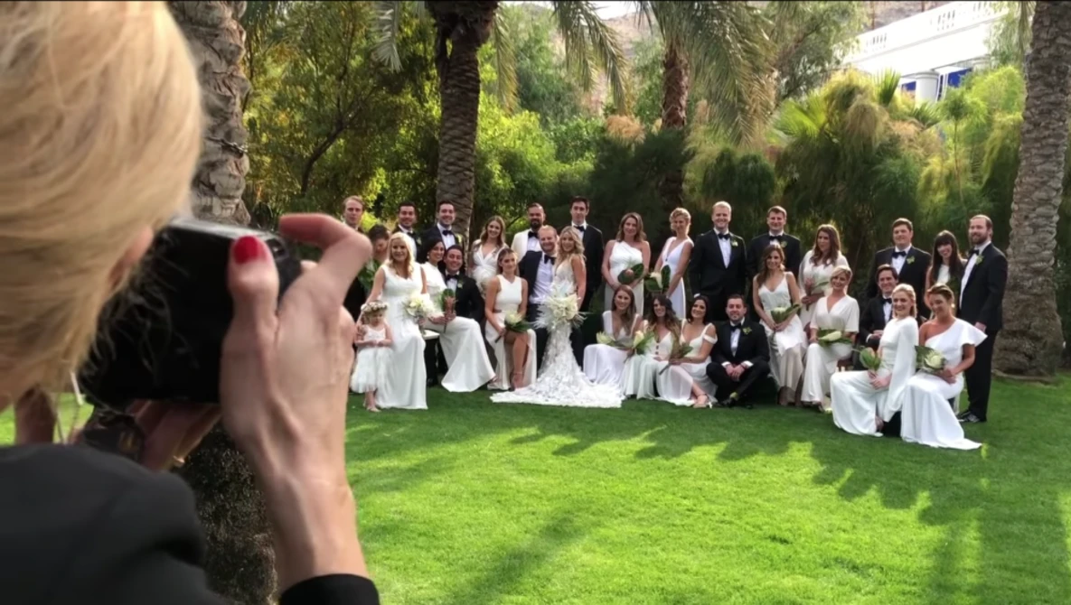 Photographer capturing a formal group portrait of wedding party on manicured lawn surrounded by palm trees.