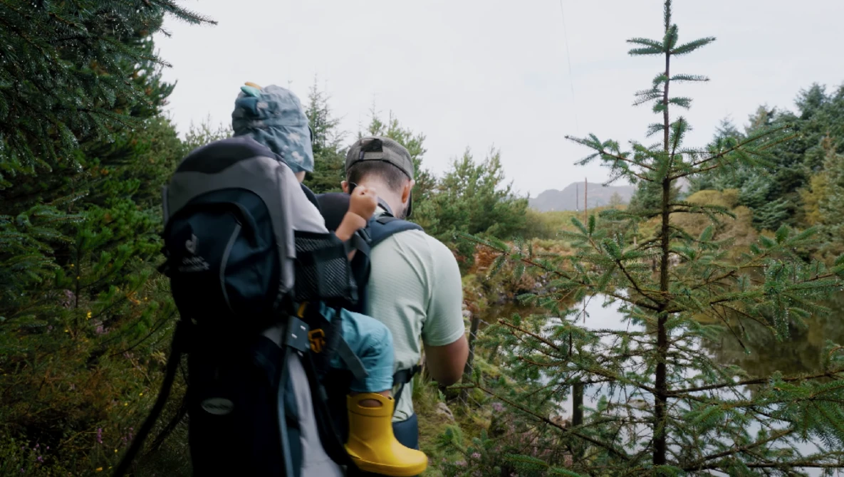 Backpacker wearing large hiking pack walks along forested trail beside calm water.
