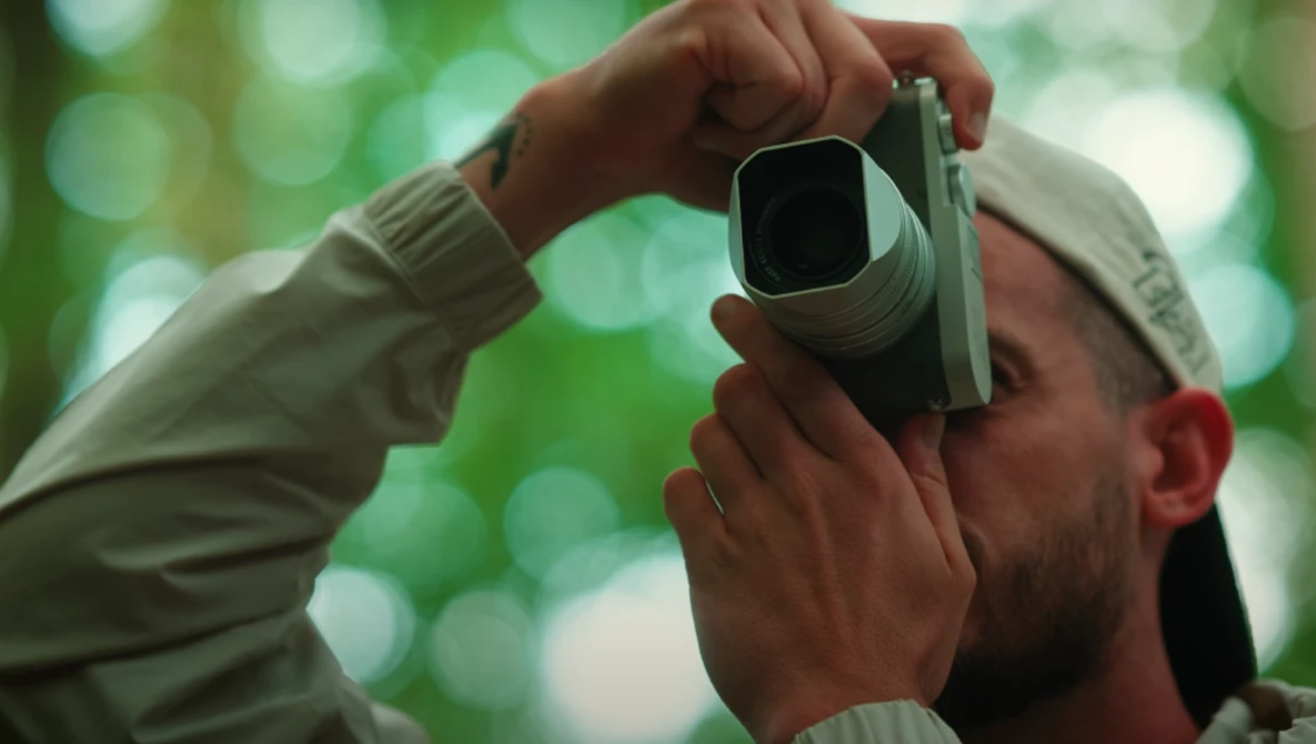 Photographer holding a rangefinder camera to their eye against a soft, blurred green foliage background.