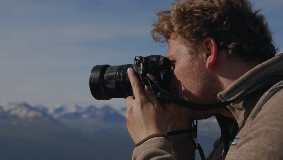 Photographer with curly reddish-brown hair holds a mirrorless camera with telephoto lens to his face, composing a shot of snow-capped mountains under clear blue sky.