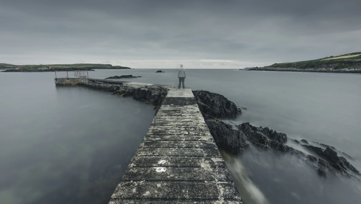 Long stone pier extending into calm gray water with rocky outcrops and distant coastline under overcast sky.