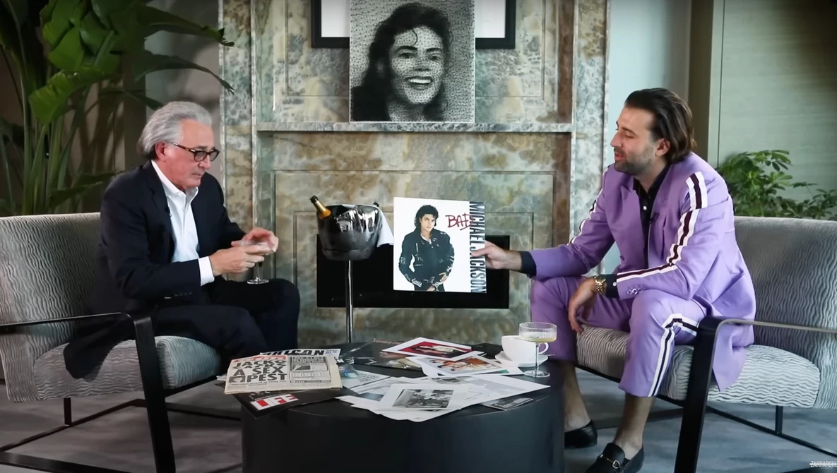 Two men seated across from each other in a studio setting with framed photographs and memorabilia on a table between them.