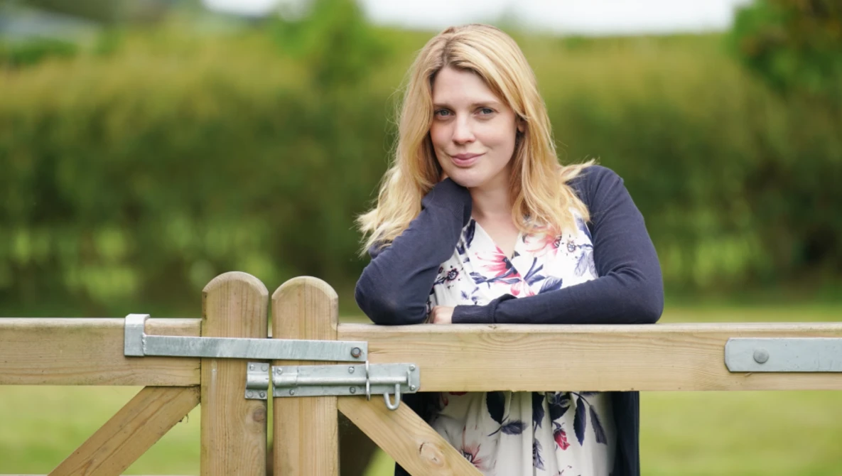 Woman with blonde hair leaning against a wooden fence in an outdoor garden setting.