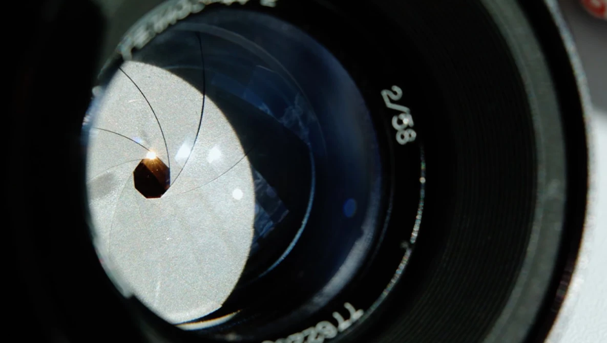 Long-legged spider viewed through a camera lens aperture showing depth of field and lens markings.