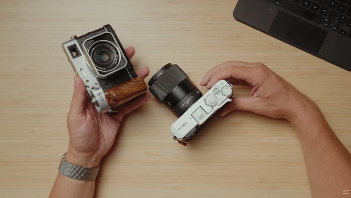 Hands holding two vintage rangefinder cameras with interchangeable lenses on a wooden desk.
