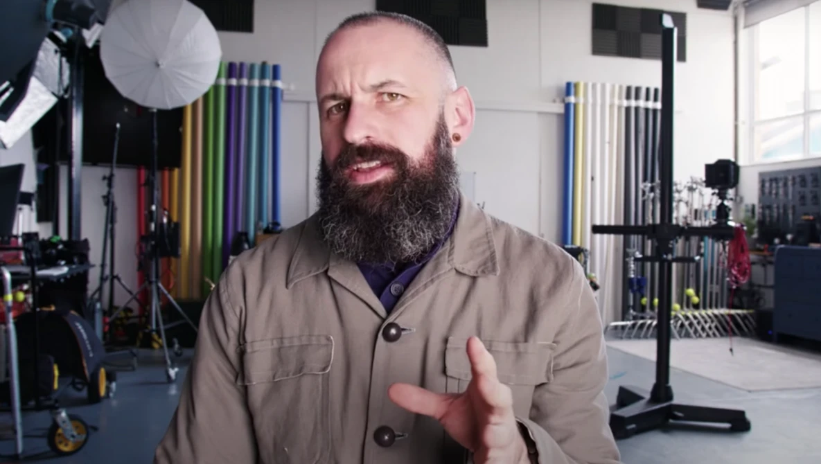 Bearded man in tan work shirt gesturing while speaking in a professional studio setting.