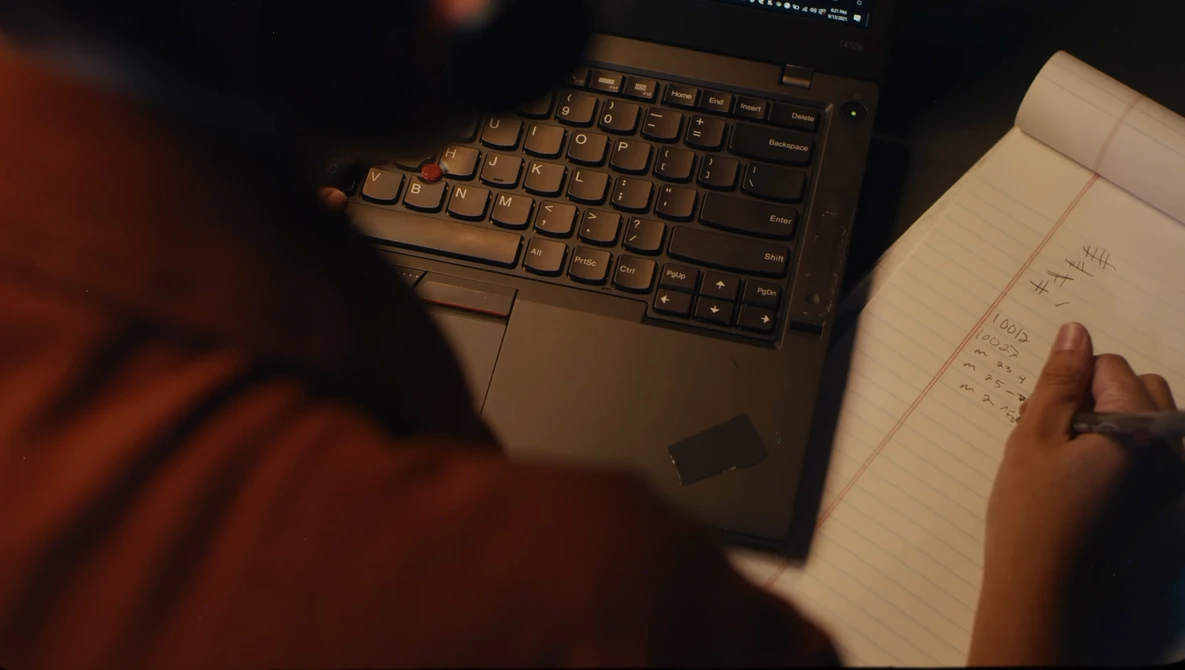 Person in rust-colored sleeve pointing at handwritten notes beside a laptop keyboard during evening work session.
