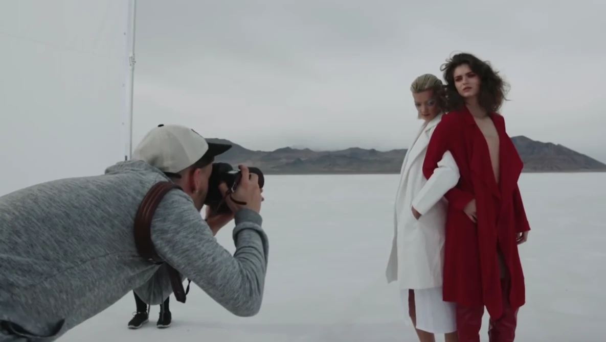 Photographer in white cap and gray jacket shooting two women in red and white clothing standing by a calm body of water.