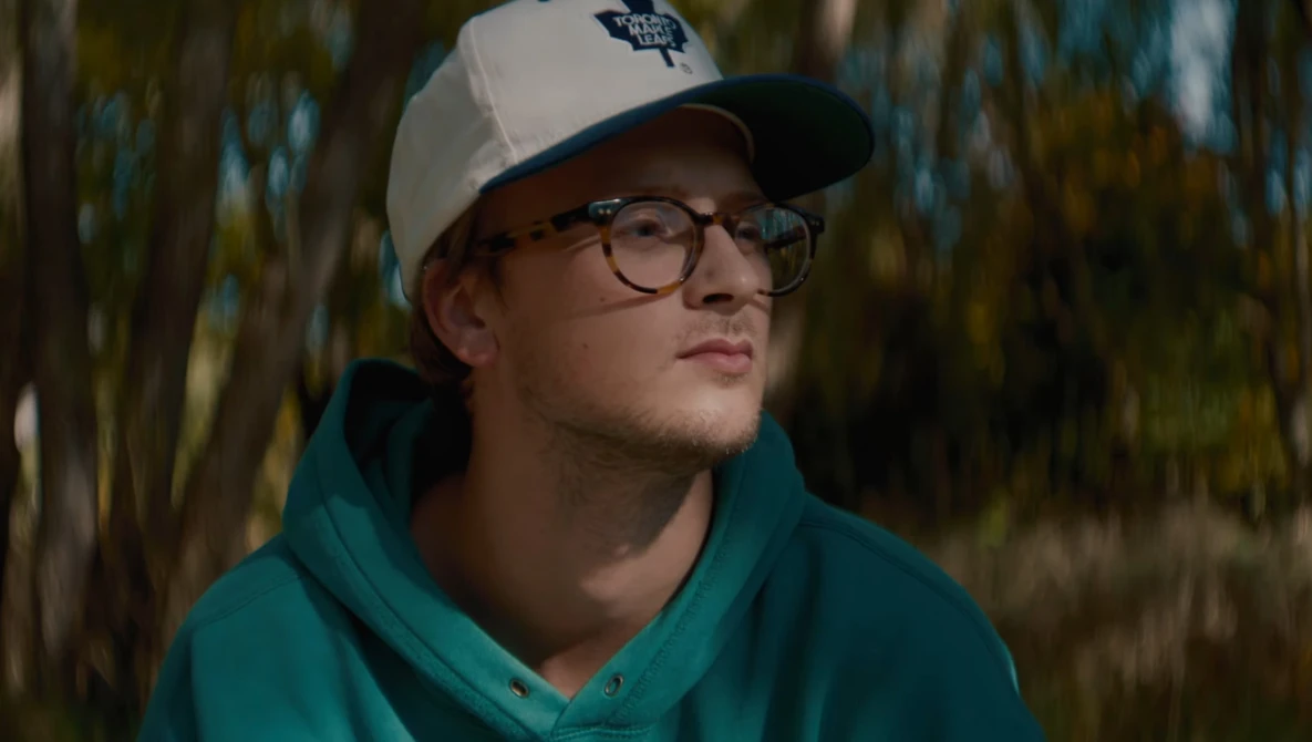 Young man wearing glasses and a teal hoodie with a white and navy baseball cap, photographed outdoors against blurred foliage.