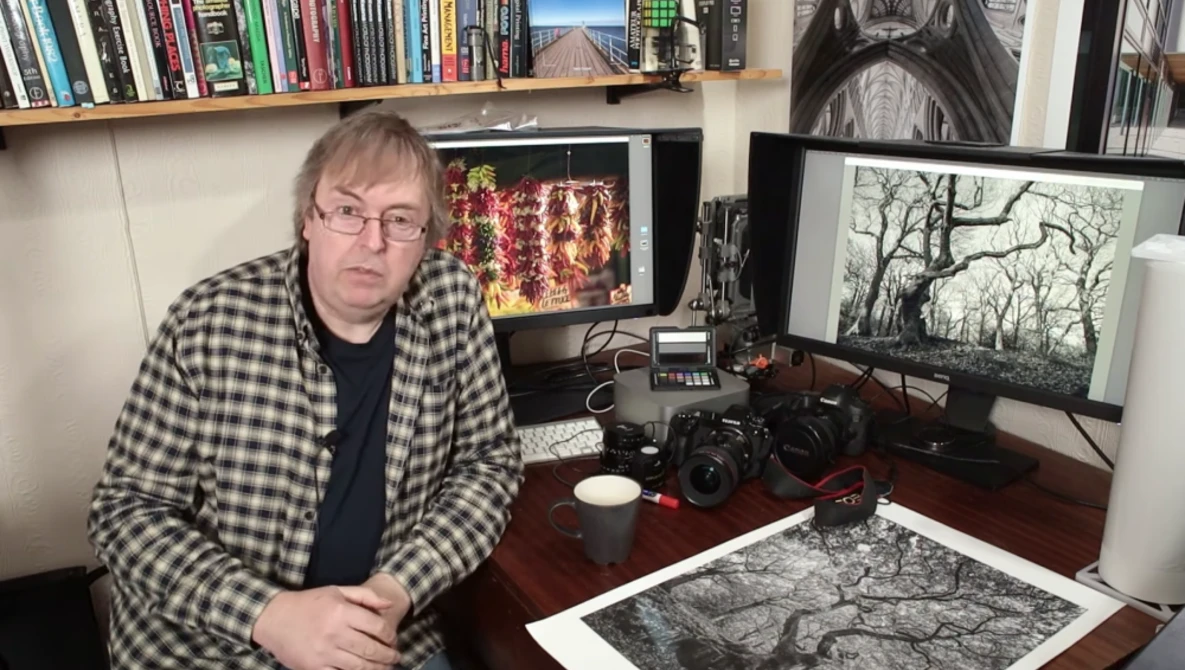 Photographer at desk surrounded by cameras, monitors displaying landscape images, and film prints.