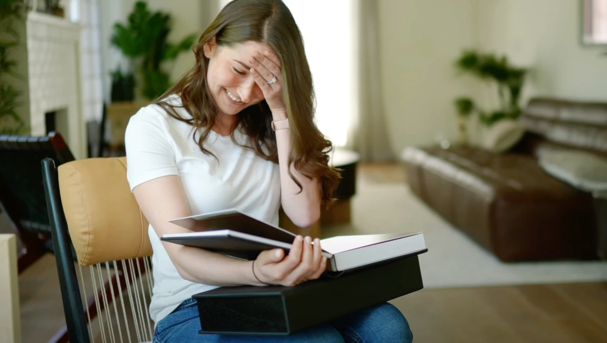 Woman sitting indoors holding stacked books with hand to forehead, appearing stressed or overwhelmed.