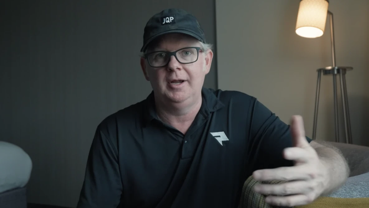 Man wearing glasses and black cap gesturing while speaking indoors.