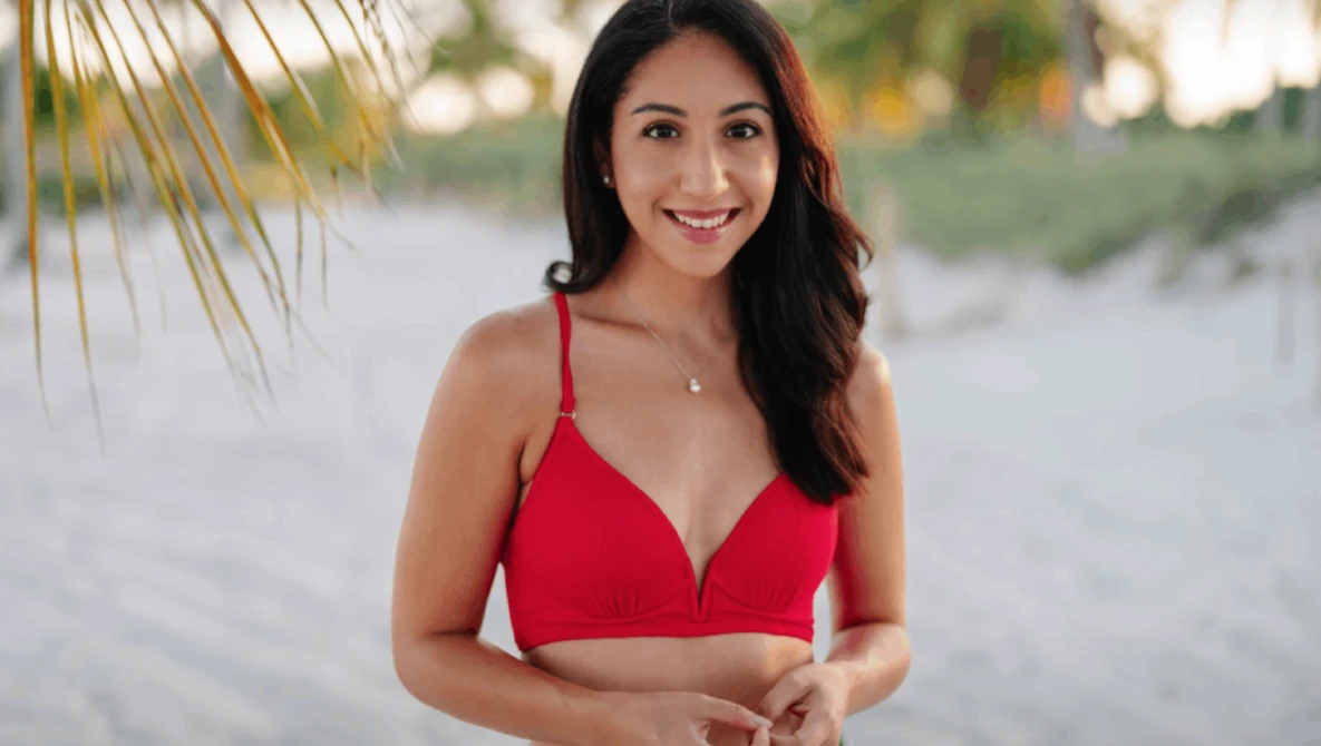 Woman in red bikini top smiling at camera on beach at golden hour with palm fronds visible.