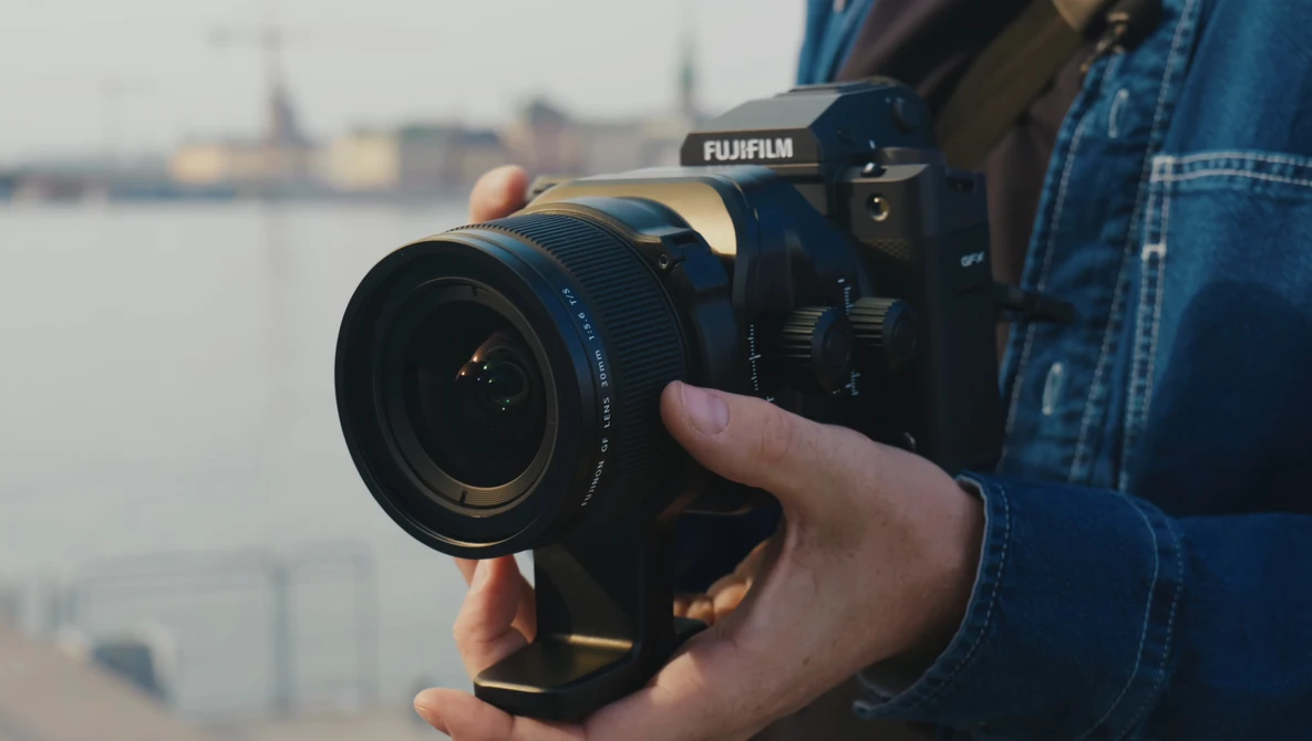 Photographer holding a Fujifilm mirrorless camera with attached lens, positioned toward a waterfront cityscape.