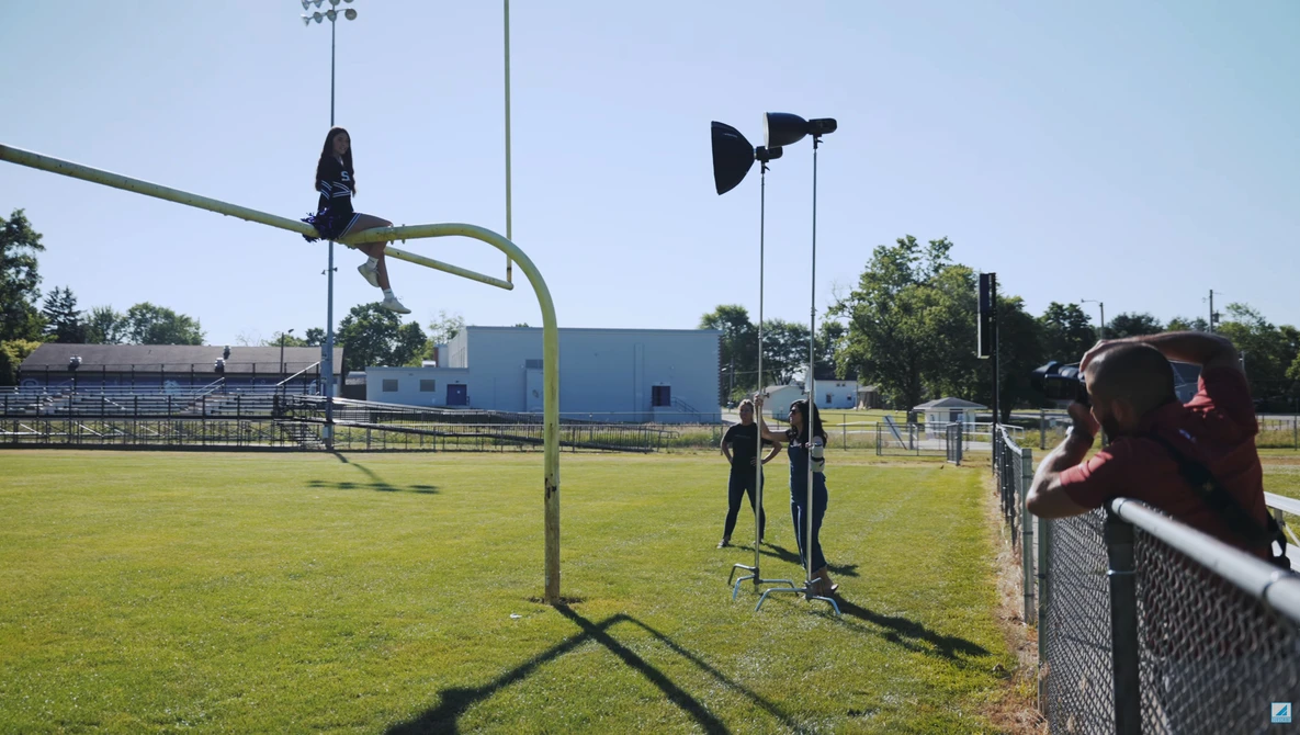 Two people standing on a sunny baseball field with tall light poles and a blue building visible in the background.