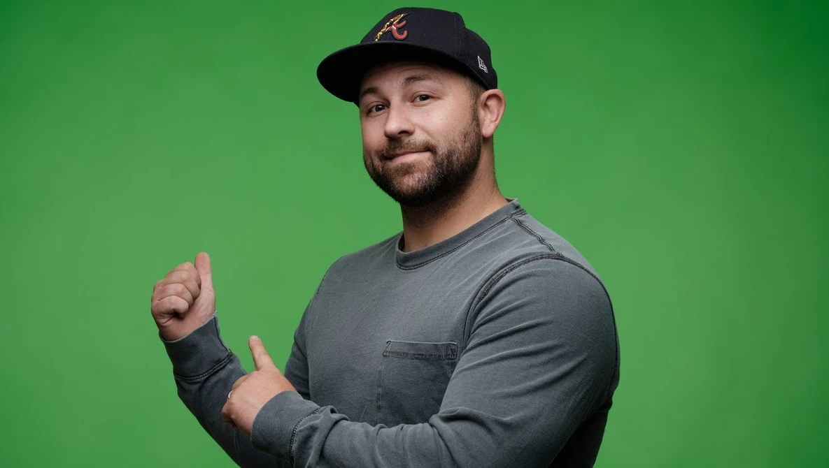 Man wearing black cap and gray long-sleeve shirt giving a thumbs-up gesture against green background.