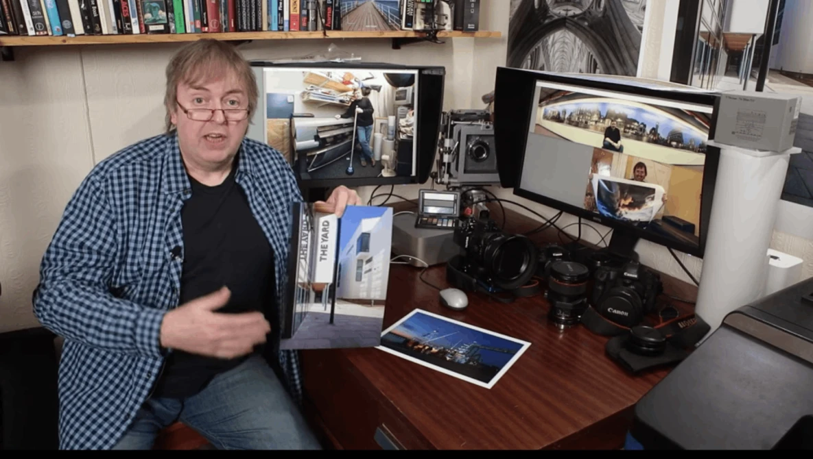 Man wearing glasses and blue plaid shirt gestures while seated at desk with photography equipment and monitors.