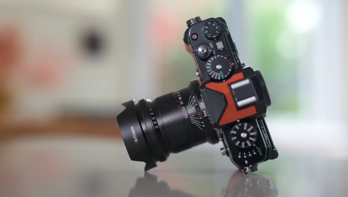 Black mirrorless camera with red accent grips and vintage manual focus lens mounted, photographed against blurred indoor background.