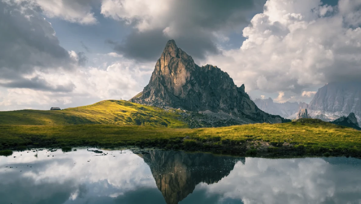 Dramatic mountain peak reflected in still water with golden meadow and cloudy sky.