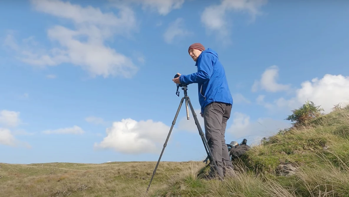 Photographer using a tripod and camera on a hillside under blue sky with scattered clouds.