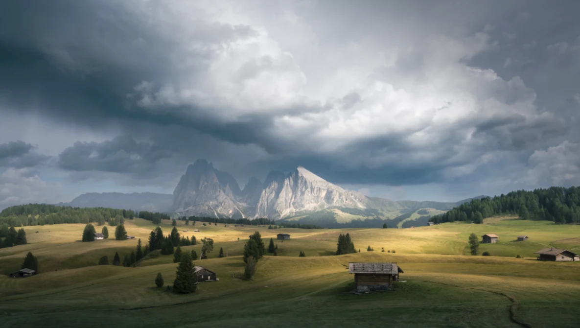 Alpine mountain valley with dramatic storm clouds, scattered farmhouses, and forested slopes beneath towering peaks.