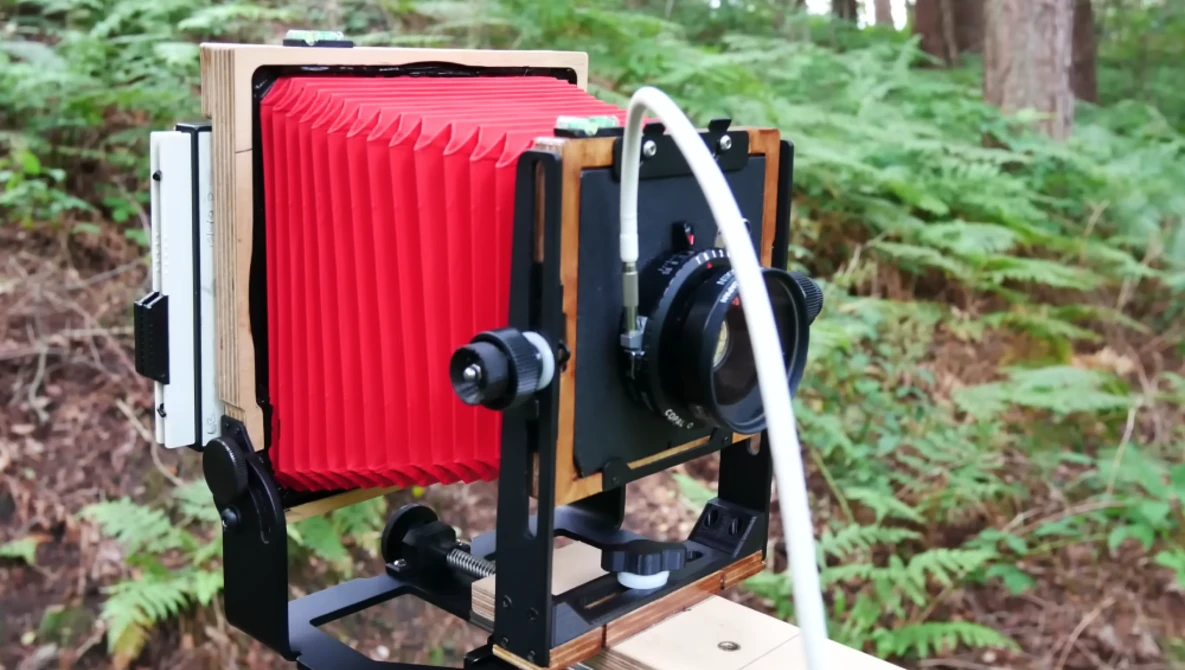Large format camera with red bellows mounted on wooden frame in forest setting.