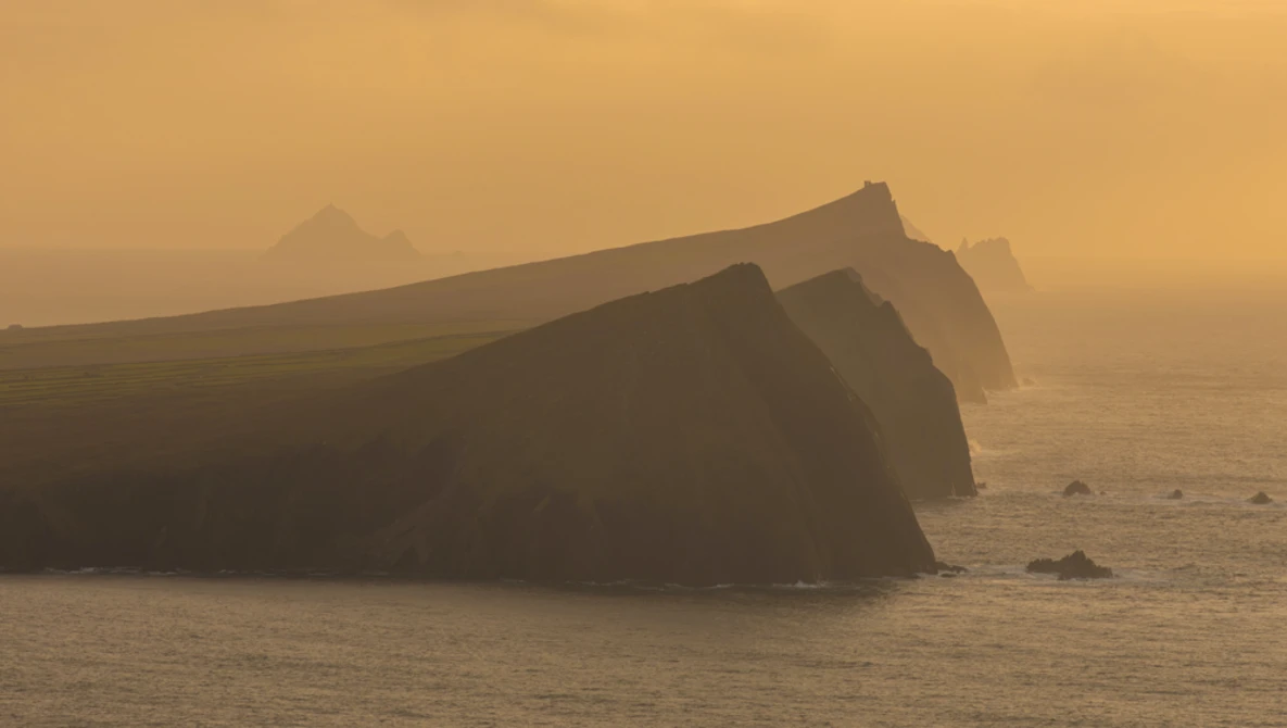 Dramatic coastal cliffs shrouded in golden mist, their steep faces rising from calm waters in atmospheric conditions.