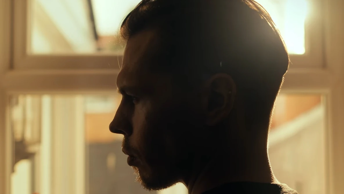 Man in profile silhouette backlit by warm window light, showing contemplative expression.