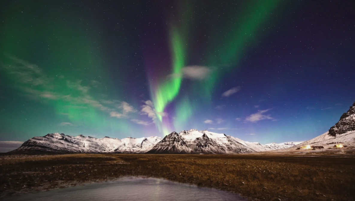 Northern lights dancing in green and purple bands above snow-capped mountains and a frozen lake.