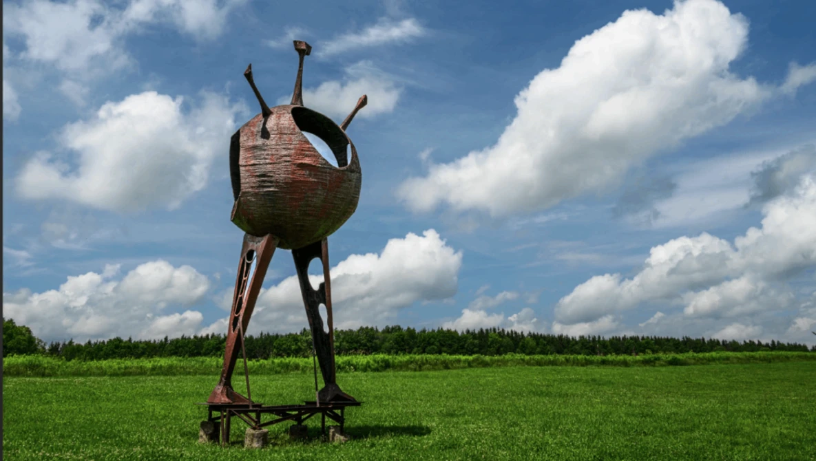 Rusted metal sculptural vessel with three protruding legs standing alone in a vast green field under cloudy blue sky.