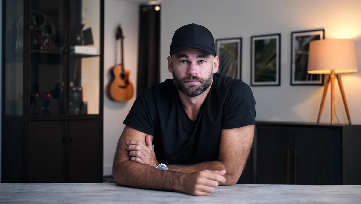 Man in black cap and shirt leaning against table in studio with warm lighting and acoustic guitar visible in background.