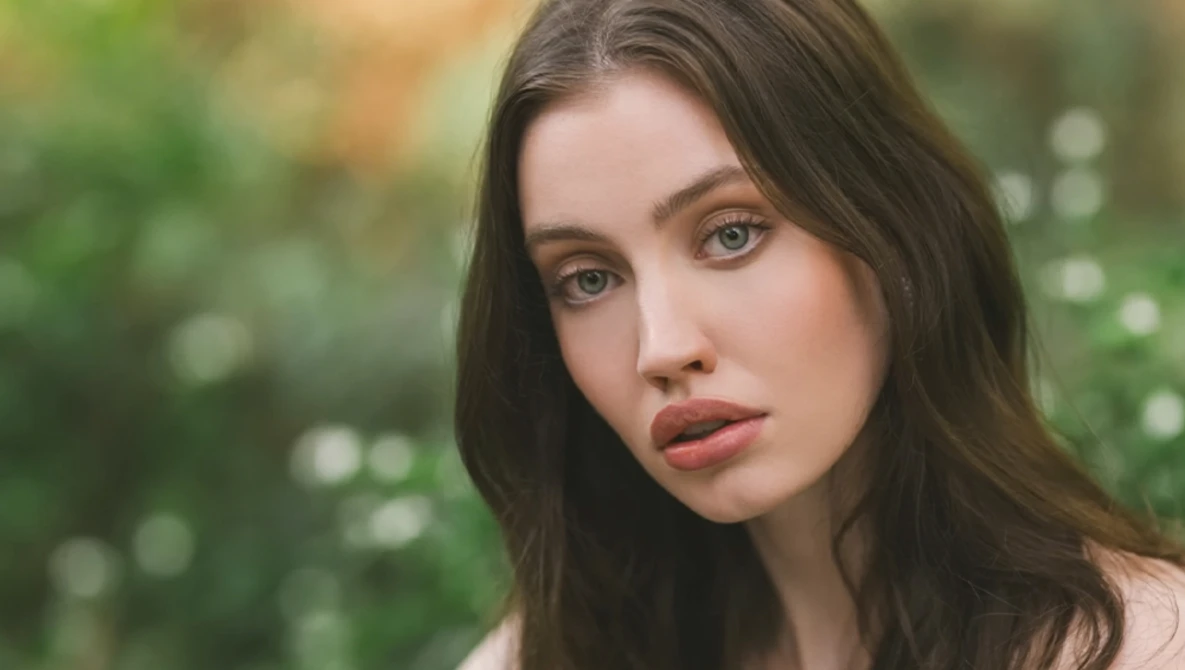 Woman with long dark hair and green eyes looking directly at camera against soft blurred green background.