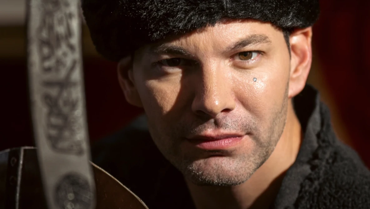 Man with dark hair and facial hair wearing a black knit beanie and dark sweater, gazing to the side with dramatic side lighting.