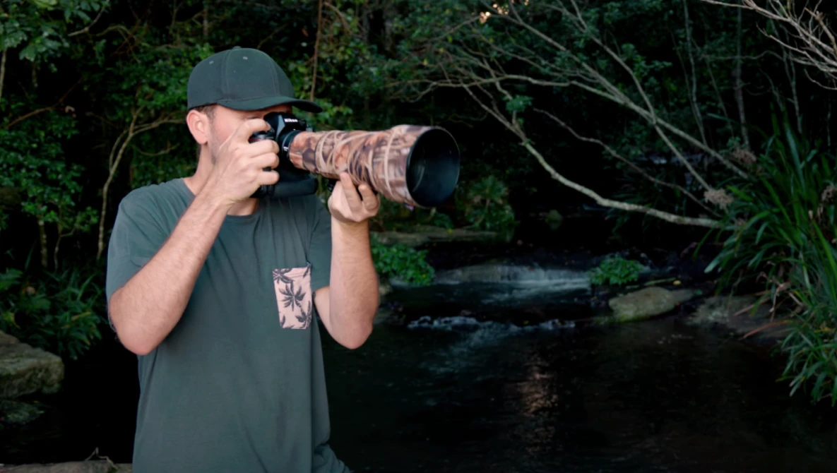 Photographer with telephoto lens in forest setting near water.