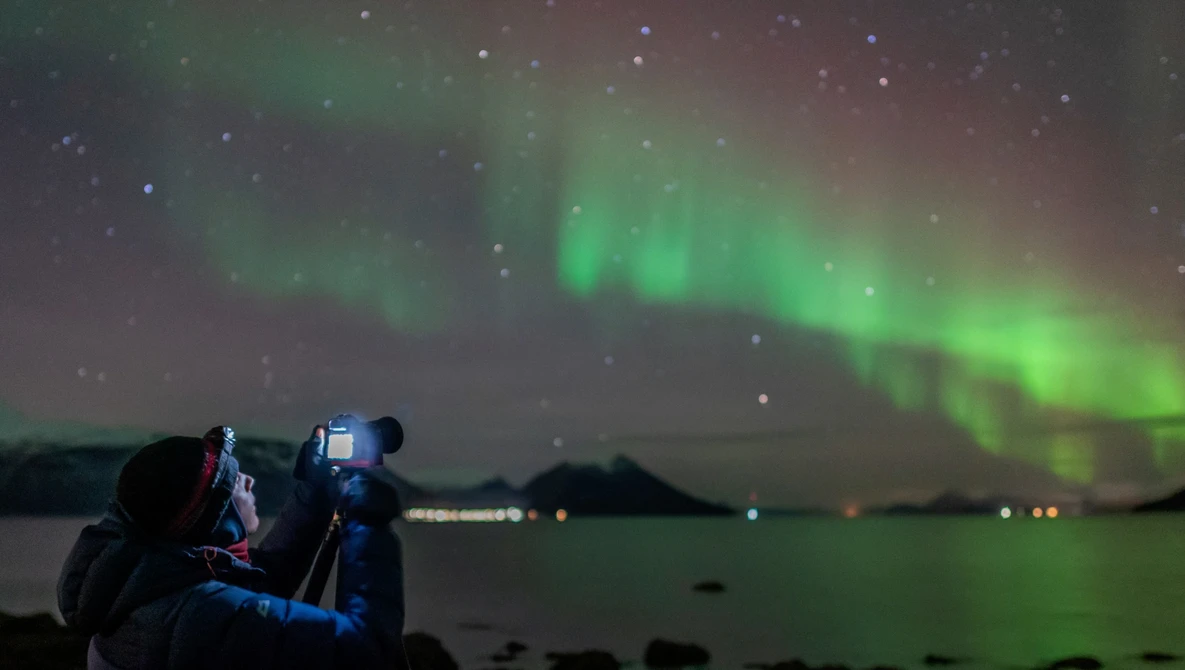 Photographer with camera on tripod capturing aurora borealis over dark water at night.