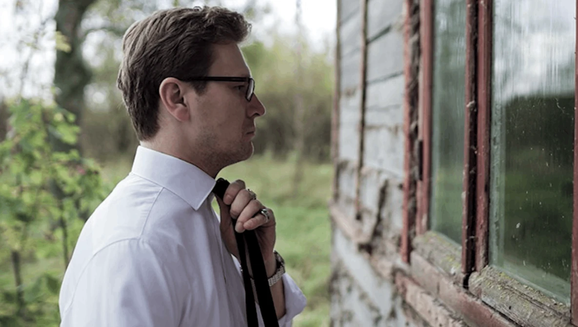 Man in white shirt and sunglasses looking toward weathered greenhouse window with green foliage backdrop.