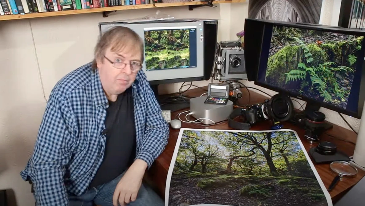 Photographer reviewing a forest landscape print at his desk workstation with dual monitors.