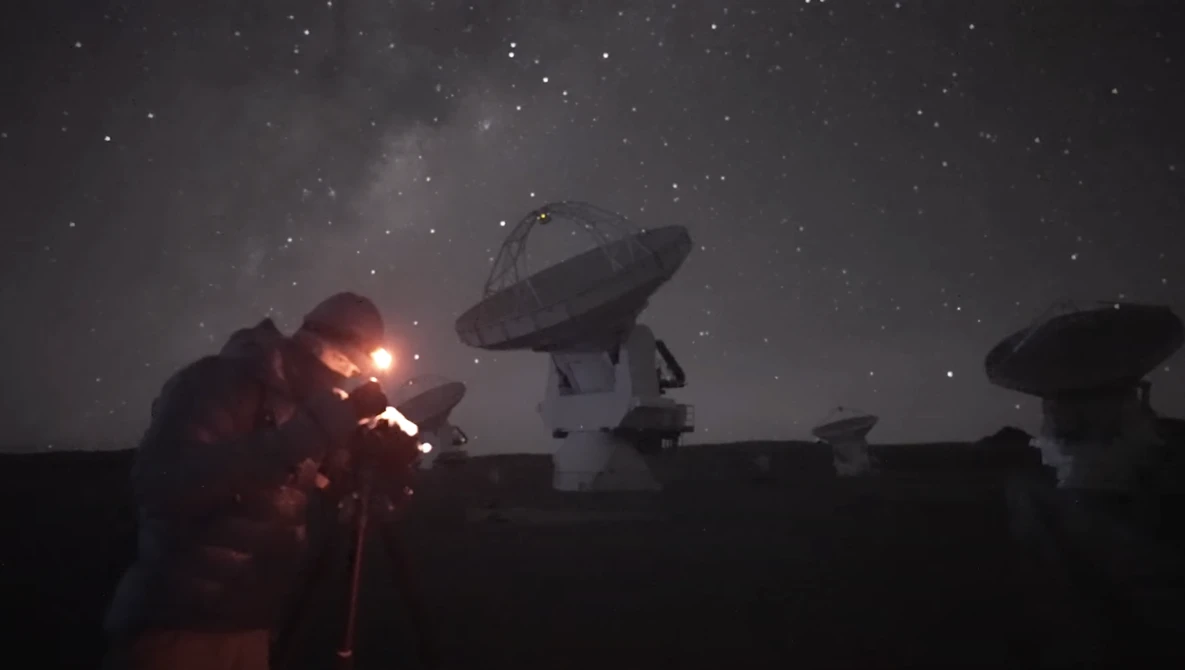 Radio telescope dishes silhouetted against a starry night sky with an explosion or bright light source illuminating one telescope in orange and red.